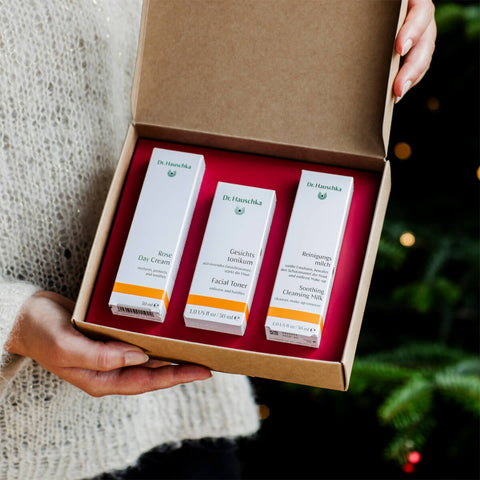 Hand holding open box of Dr Hauschka Rose Ritual Christmas Set, showing the products inside. A 3-step morning care ritual box, featuring deluxe sample sizes of Soothing Cleansing Milk, Facial Toner, and a full size tube of Rose Day Cream. The box design is elegant and minimalistic, showcasing the brand's signature natural skincare products.