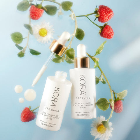 White dropper bottle of KORA Organics Silky Sun Drops Protection Serum (30ml) styled with raspberries and flowers, a certified organic SPF15 serum that protects skin from aging, blue light, UV damage, and pollution while hydrating without leaving a white cast.