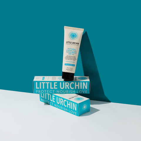 100ml bottle of Little Urchin Natural Clear Zinc Sunscreen SPF 30 balanced on a stack of its boxes on a blue surface. The sunscreen is a hydrating, vegan formula that provides UVA and UVN broad spectrum protection from the sun while also being 4 hours water-resistant. Packaged in a cream tube with minimal blue and black text on the label.