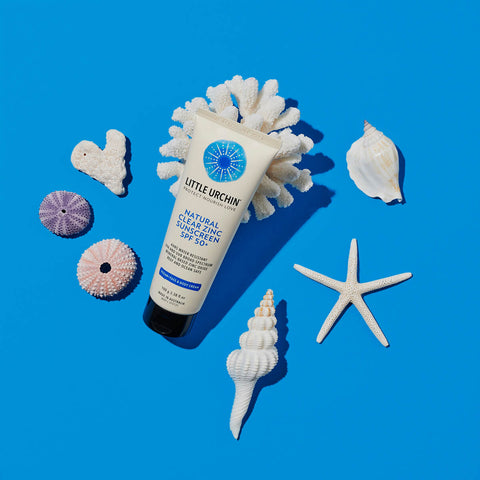 100ml bottle of Little Urchin Natural Clear Zinc Sunscreen SPF 50+ on a blue surface with coral and seashells scattered around the scene. The sunscreen is a hydrating, vegan formula that provides UVA and UVN broad spectrum protection from the sun while also being 4 hours water-resistant. Packaged in a cream tube with minimal blue and black text on the label.