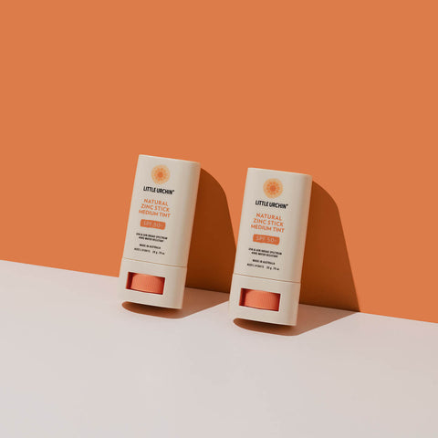 20g tube of Natural Zinc Stick SPF 50+, a medium tint, hydrating, vegan formula that provides UVA and UVB broad-spectrum protection from the sun while also being 4 hours water-resistant, placed against an orange background. Packaged in a cream tube with minimal orange and black text on the label.