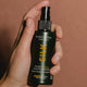 Hand holding 90ml bottle of Madara Silk Micro-Keratin Healthy Hair Mist, a natural certified micro-keratin spray that helps repair fibre damage, reduce breakage and split ends. Packaged in a dark green bottle.