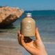 Hand holding bottle of cleansing powder with rosalina and marshmallow in front of a beach.