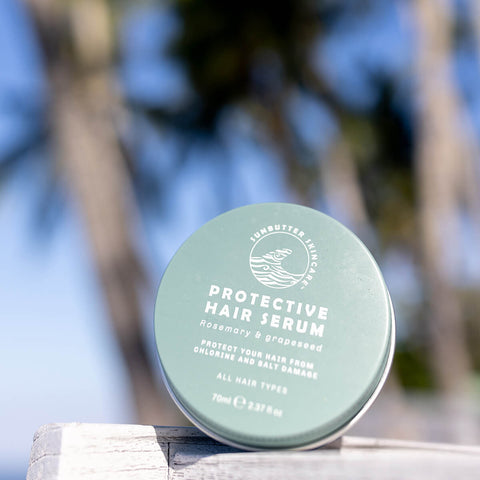 70ml tin of SunButter Oceans Protective Hair Serum with rosemary and grapeseed, set against a blurred background of palm trees. The blue tin features simple white text and is designed to nourish and protect hair from chlorine and salt damage. Suitable for all hair types.