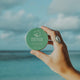 A hand holding a 70ml tin of SunButter Oceans Protective Hair Serum with rosemary and grapeseed. The tin is set against a blurred background of the ocean. The blue tin, featuring simple white text, is designed to nourish and protect hair from chlorine and salt damage. Suitable for all hair types.