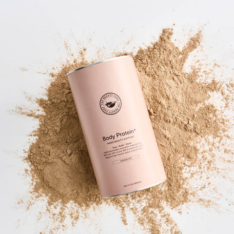 Light brown coloured 600g tin of The Beauty Chef Body Protein+ Inner Beauty Support in chocolate flavour on top of powder.