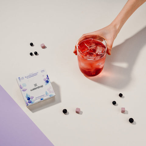 A hand holding a glass of blackcurrant and elderberry flavoured water with ice, next to a white box of 12 waterdrop Microdrink cubes in the Boost flavour. The box displays a blackcurrant and elderberry flavour, with purple and blue berry illustrations on the front. Scattered around the scene are small hydration cubes and berries on a light cream surface.