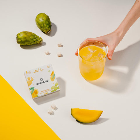 A hand holding a glass of mango and cactus fruit flavoured water with ice, next to a white box of 12 waterdrop Microdrink cubes in the Glow flavour. The box displays a mango and cactus fruit flavour, with yellow mango and cactus illustrations on the front. Scattered around the scene are small hydration cubes, a slice of mango and cactus fruit on a light cream surface.