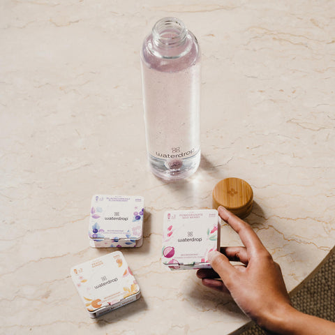 A hand holding a box of waterdrop Microdrink cubes in the Love flavour, featuring pomegranate and goji berry. A box of waterdrop Microdrink cubes in the Peach and Youth flavour lay on a cream coloured, marble surface next to a waterdrop branded reusable glass bottle with a Microdrink cube dissolving in the water.