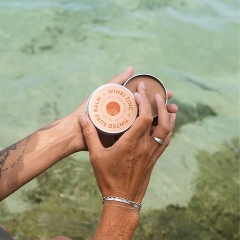 Hand holding 70g tin of Winki Zinc Tinted Surf Balm, a reef safe and water resistant zinc made with natural ingredients, including beeswax and cocoa butter.